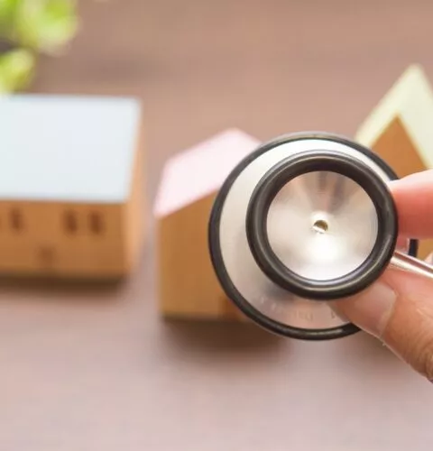 Une main tenant un stéthoscope pointé vers des maisons miniatures en bois, symbolisant le diagnostic immobilier
