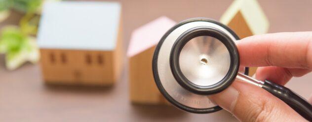Une main tenant un stéthoscope pointé vers des maisons miniatures en bois, symbolisant le diagnostic immobilier